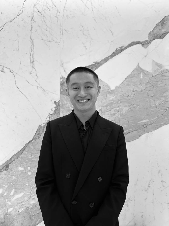 Black and white portrait of Ethan Legarta, smiling, in a dark double-breasted suit in front of a marble wall